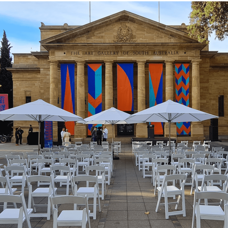 The front facade of the Art Gallery of South Australia stands in elegant contrast to a contemporary outdoor event setup. Beneath the gallery’s classical columns and vibrant geometric banners, rows of crisp white chairs and umbrellas—supplied by Australian Hiring Company—create a welcoming, structured space for guests. The arrangement suggests a formal gathering, blending cultural sophistication with modern event styling. A few attendees converse near the entrance, while the shaded seating offers comfort and cohesion, enhancing the gallery’s role as both an artistic and civic venue.