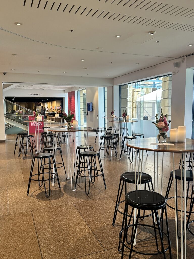 Inside the Art Gallery of South Australia, a contemporary event space comes to life with hairpin-leg tables and stools supplied by Australian Hiring Company. Each table is styled with minimalist floral arrangements and pillar candles, creating an inviting, modern ambiance. The setting is complemented by catering from _eats, part of the 2KW Group, whose presence adds a refined culinary touch to the gallery’s vibrant cultural atmosphere. Natural light floods the space, enhancing the clean lines and thoughtful design—ideal for intimate gatherings, exhibition launches, or donor receptions.