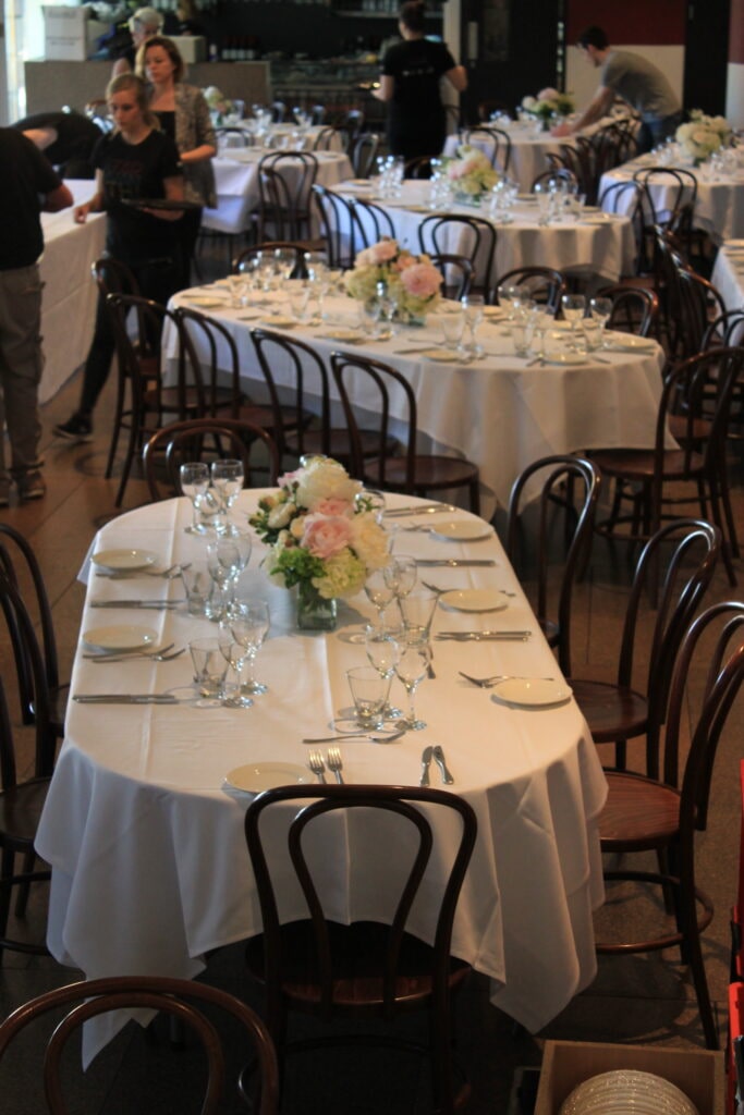 An elegant event unfolds inside the Art Gallery of South Australia, where oval tables dressed in crisp white linens are paired with classic bentwood chairs—furniture and tableware provided by Australian Hiring Company. Each table is meticulously set with polished glassware, silver cutlery, and white ceramic plates, complemented by soft floral centerpieces in blush and ivory tones. The refined dining experience is elevated by catering from _eats, part of the 2KW Group, whose culinary presentation matches the gallery’s sophisticated ambiance. The overall setting blends heritage architecture with contemporary hospitality, ideal for gala dinners, cultural receptions, or donor events.