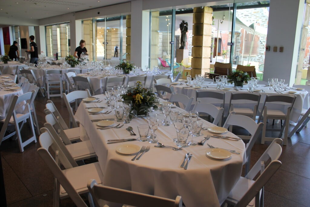 Inside the Art Gallery of South Australia, a sophisticated event setup features oval tables dressed in white linen and surrounded by classic white Americana chairs—furniture and tableware provided by Australian Hiring Company. Each table is elegantly set with polished silverware, glassware, and white ceramic plates, complemented by fresh floral centerpieces in soft yellow and green tones. Natural light pours through the gallery’s expansive windows, illuminating the space and highlighting the architectural details beyond. Catering by _eats from the 2KW Group adds a contemporary culinary touch, completing a seamless blend of heritage, hospitality, and design.