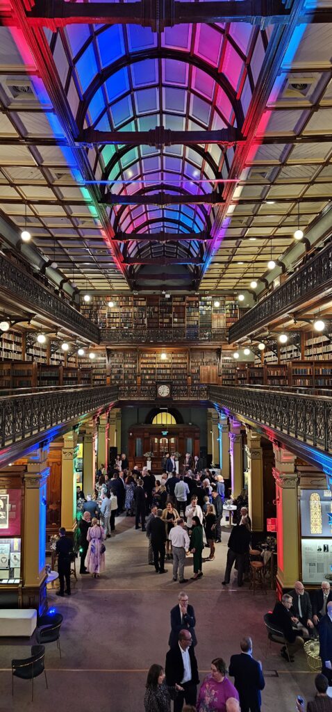 Cocktail-style wedding reception hosted by Australian Hiring Company in the iconic Mortlock Library, Adelaide. The grand heritage space glows with ambient red, blue, and purple lighting, casting a modern elegance across iron balconies, classical columns, and multi-tiered bookcases. Guests mingle in formal attire beneath the soaring arched ceiling, surrounded by curated lounge furniture and cocktail tables supplied by Australian Hiring Company. The setup balances historic grandeur with contemporary flair, showcasing expert event management and premium furnishings in one of South Australia’s most treasured cultural venues.