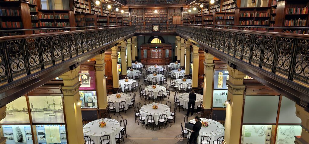 Elegant dining setup amid towering bookcases and historic ironwork inside Mortlock Chamber, State Library of South Australia. Round tables with floral centerpieces and white tablecloths arranged beneath ornate balconies and antique clock. Heritage-listed venue available for weddings and private events through Australian Hiring Company.