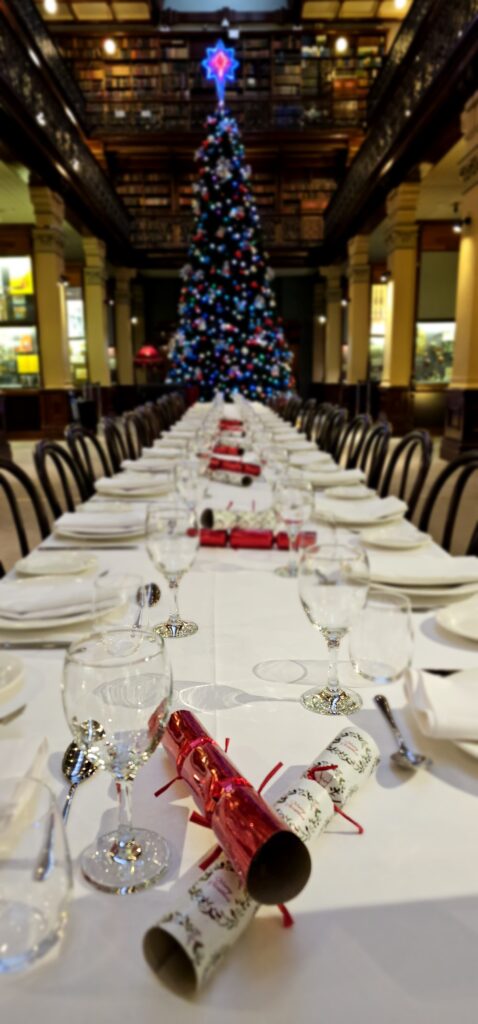 Christmas function hosted by Australian Hiring Company in the historic Mortlock Library, Adelaide. A long banquet table stretches through the grand hall, elegantly dressed in white linens and set with fine glassware, silver cutlery, and festive red-and-white Christmas crackers. A towering, ornamented Christmas tree glows in the background, adding warmth and seasonal charm beneath the venue’s soaring ceilings and classical columns. Surrounded by ornate balconies and heritage bookcases, the scene showcases Australian Hiring Company’s premium furniture and expert event management, transforming this cultural landmark into a refined celebration space.
