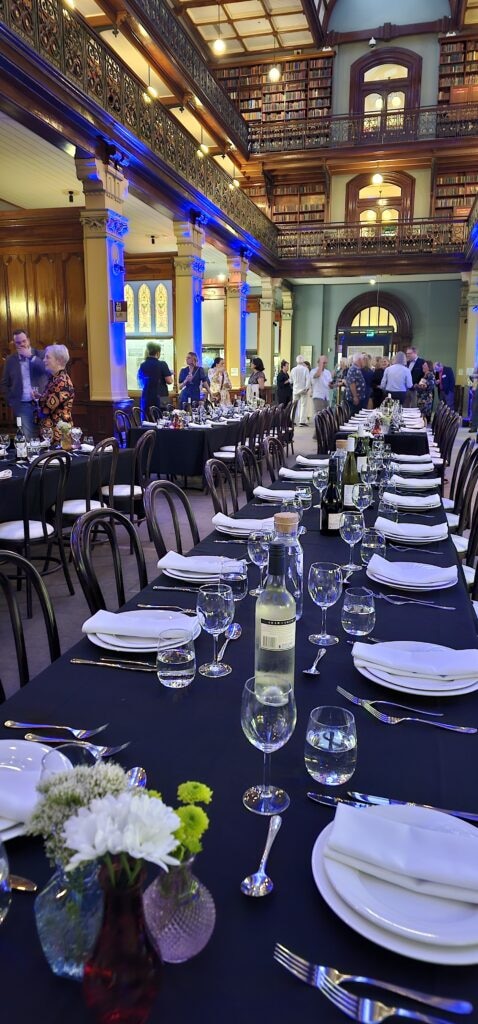 Long banquet setup for Law Australasia inside Mortlock Chamber, State Library of South Australia. Black tablecloths, white dinnerware, and floral centerpieces arranged beneath vaulted ceilings, stained glass windows, and heritage ironwork. Available for corporate events and private functions through Australian Hiring Company.