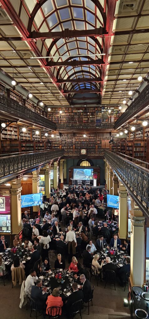 Corporate dinner for CommSec inside Mortlock Chamber, State Library of South Australia. Guests seated at round tables beneath vaulted glass ceilings and heritage bookcases, with branded screens and floral centerpieces enhancing the elegant event setting. Available for private functions and business events through Australian Hiring Company.