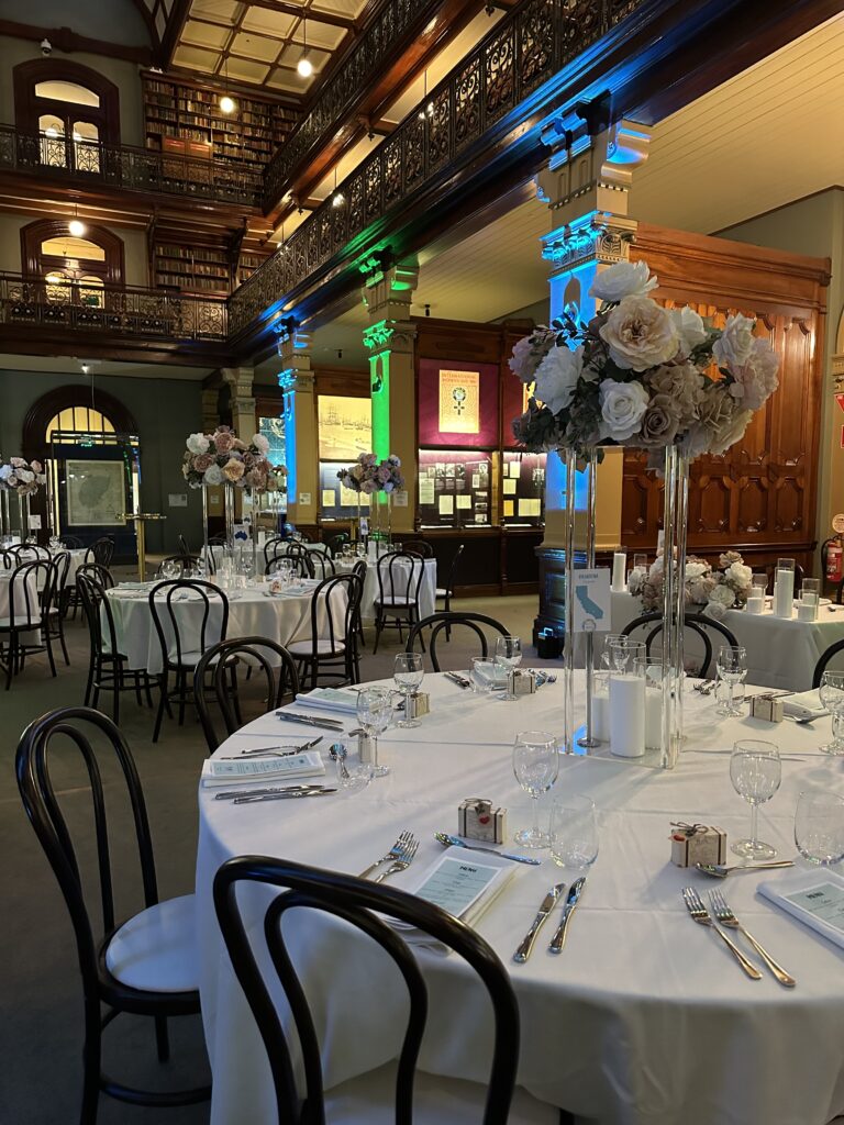  Round tables with floral centerpieces and guest settings inside Mortlock Chamber, State Library of South Australia. Heritage-listed venue featuring historic architecture, ambient lighting, and elegant event styling—available for weddings and private functions through Australian Hiring Company.