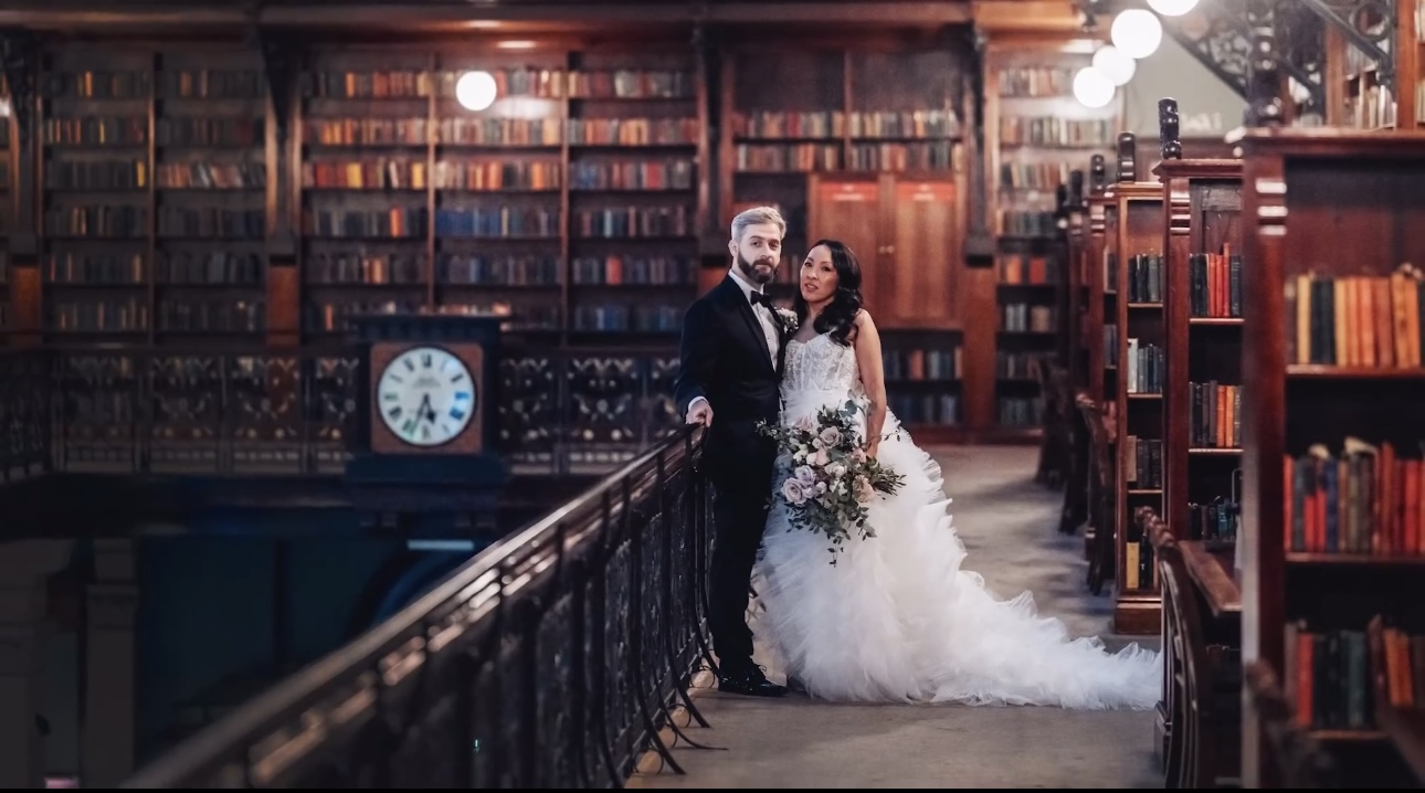 Elegant wedding couple standing on the upper balcony of Mortlock Chamber, State Library of South Australia. Bride in flowing white gown and groom in black tuxedo pose beneath historic clock and towering bookcases. A romantic, heritage-listed venue available for weddings and private events through Australian Hiring Company.