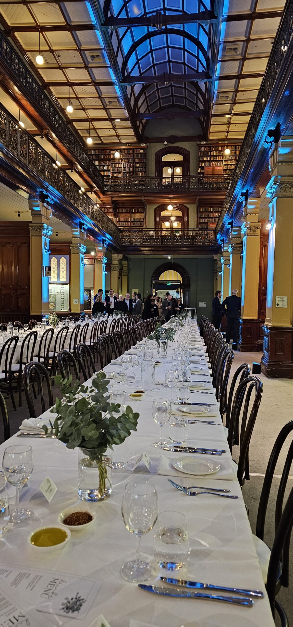 Long banquet setup with white tablecloths, greenery centerpieces, and formal guest seating inside Mortlock Chamber, State Library of South Australia. Surrounded by heritage architecture, ornate balconies, and ambient lighting. Available for weddings and private events through Australian Hiring Company.