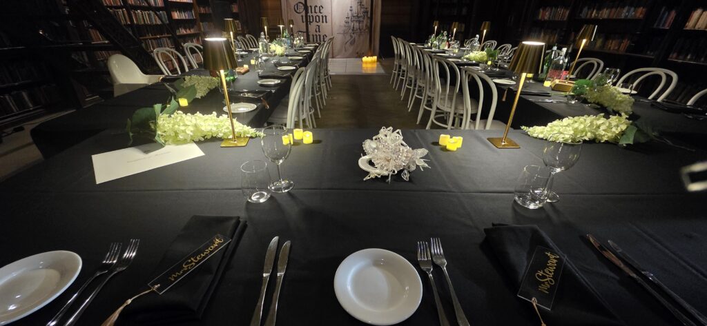 Elegant library-themed dinner event styled by Australian Hiring Company for Party Hire Adelaide, featuring black tablecloths, white bentwood chairs, gold lamps, floral centerpieces, and a “Once Upon a Time” backdrop.