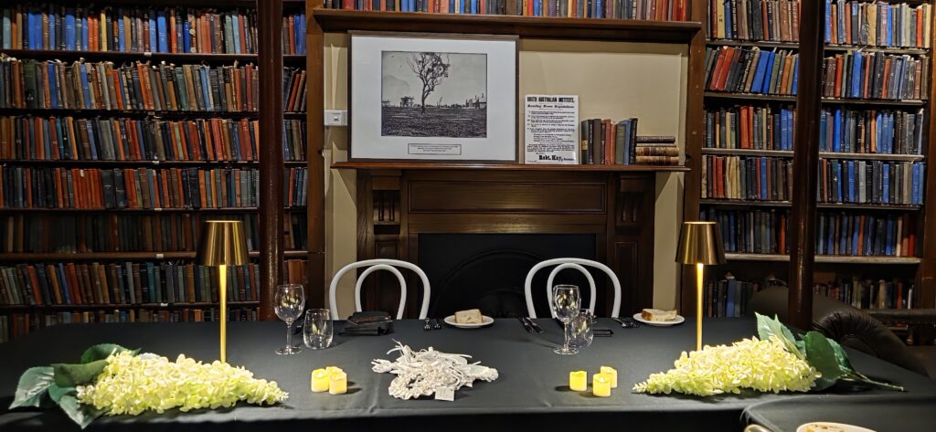 Luxurious library event setup by Australian Hiring Company for Party Hire Adelaide, featuring black tablecloths, white bentwood chairs, gold lamps, floral centerpieces, and a fireplace backdrop surrounded by towering bookshelves.
