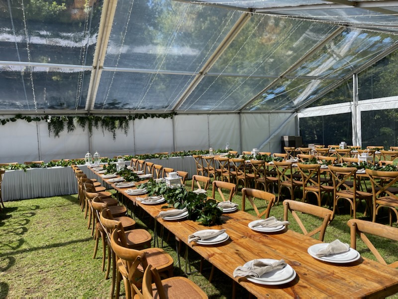 Clear-roof wedding marquee with long wooden tables, woven‑seat chairs, and green foliage runners, fully supplied and installed by Australian Hiring Company in Adelaide.