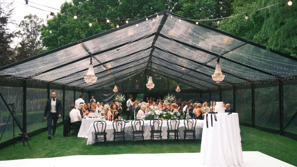Elegant outdoor dining setup under a clear marquee featuring black chairs, white tablecloths, floral centerpieces, chandeliers, and string lights — ideal for party hire, wedding hire, furniture hire, and lighting hire in Adelaide.